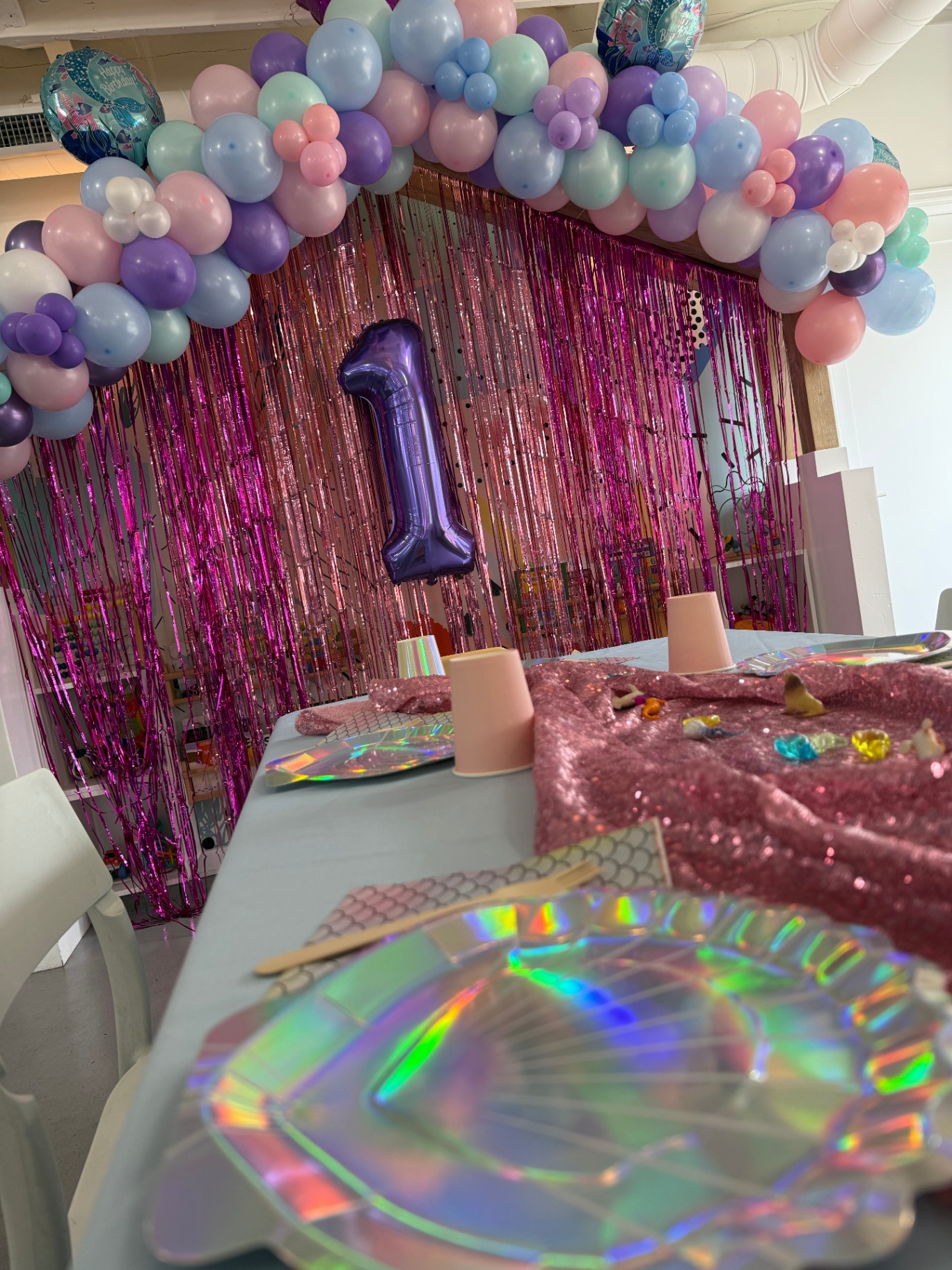 Mermaid themed first birthday at tumble HAUS with a pastel balloon arch, pink tinsel backdrop and iridescent shell place settings
