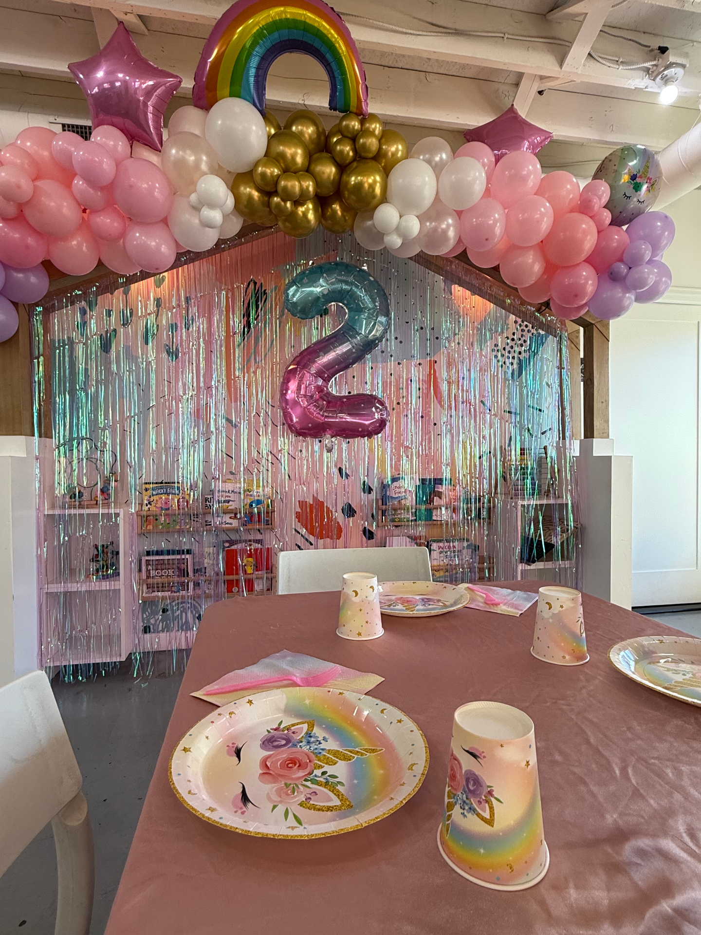 Unicorn themed 2nd birthday party at tumble HAUS with a rainbow balloon topper, pink garland, iridescent fringe backdrop and pastel unicorn tableware